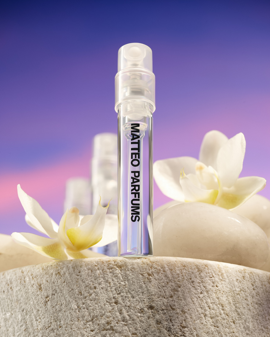 Vial of MATTEO PARFUMS luxury and prestige fine fragrance, Lovers' Dew, on a marble sea rock, with white orchid flowers, white smooth stones, and a beautiful sunset sky