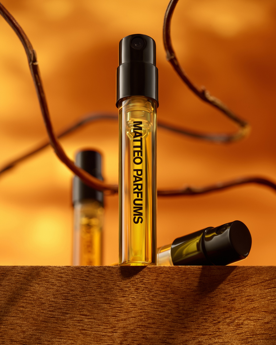 Vial of MATTEO PARFUMS luxury and prestige fine fragrance, Celadawn, on a wooden ledge, with white weaving vanilla bean pods and wood sticks, set against a golden background