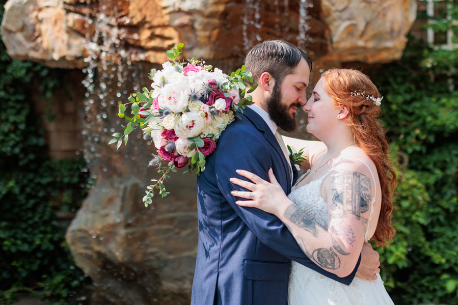 Bride and Groom in front of waterfall at the Mansion at Bald Hill. She has natural red hair and  is holding a bouquet of white and pink flowers and has an arm of tattoos. He has a beard and blue suit. 