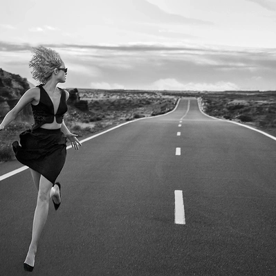 Black and white photo from a woman in a black dress sprints down a long, empty highway, the horizon stretching out before her, a symbol of endless possibilities.