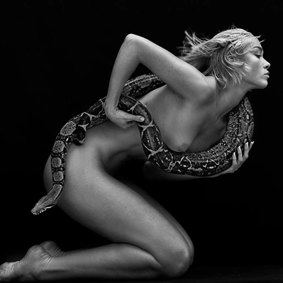 Black and white photo from a nude woman,  cradles a snake, its scales shimmering in the dark.