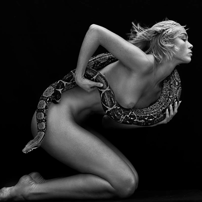 Black and white photo from a nude woman,  cradles a snake, its scales shimmering in the dark.