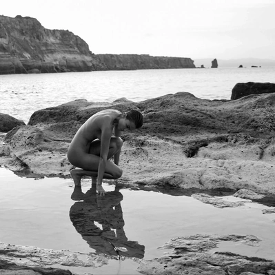 Black and white photo from coastal solitude; a nude man contemplates the ocean's edge, his reflection mirroring the serene scene.