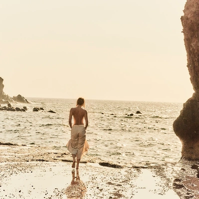 Coastal serenity, a figure in a flowing skirt, bathed in golden light on a beach with a rock on the right.