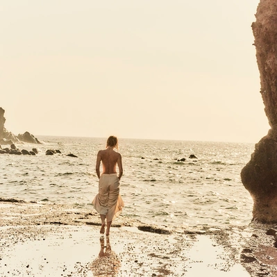 Coastal serenity, a figure in a flowing skirt, bathed in golden light on a beach with a rock on the right.