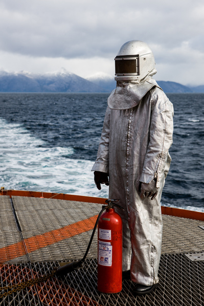 A man in a fire suit stands in the deck of a moving vessel with an extinguisher in front of his legs. 