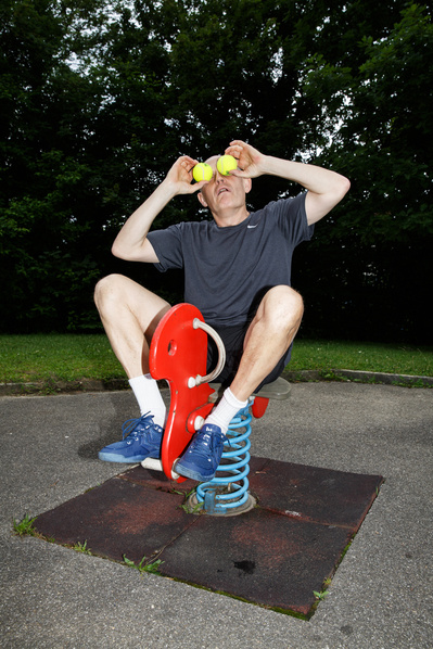 A man in shorts sits on a kids playground holding two tennis balls in his hands 