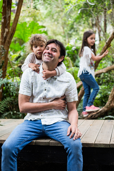 A man sitting on a wooden path is being hugged from behind by a  small girl, while there is another girl climbing a tree on the background. 