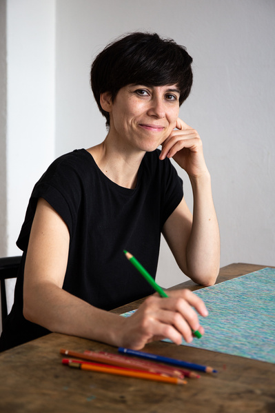 A woman sitting on a table with a drawing in front of her,  looks to the front while holding a color pencil in her hands 