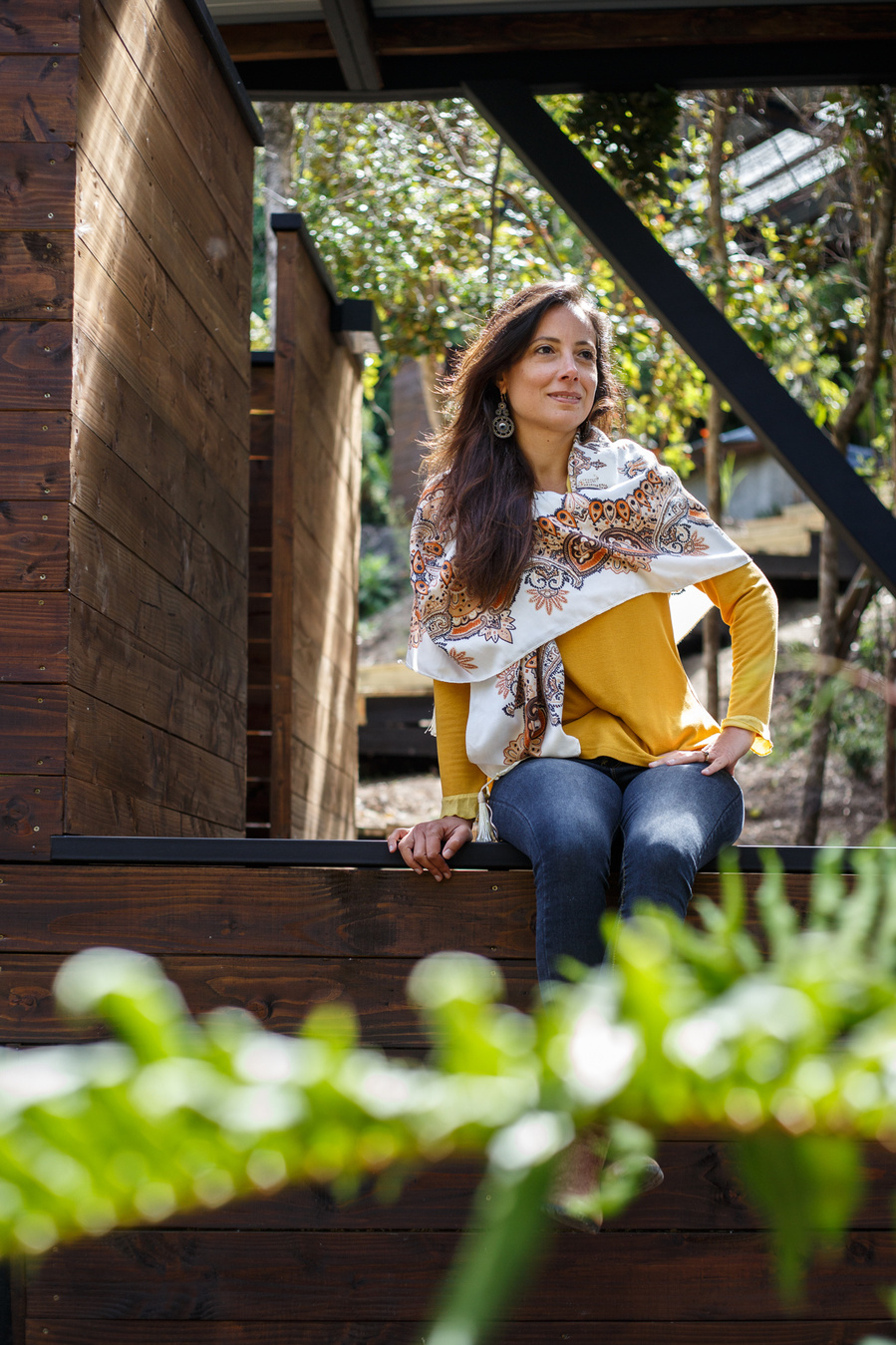 A woman with a yellow sweater sits on a wooden structures and there are blurry plants in the foreground. 