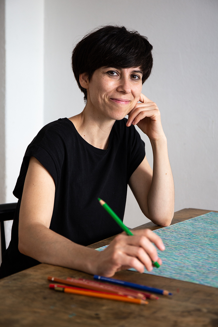 A woman sitting on a table with a drawing in front of her,  looks to the front while holding a color pencil in her hands 