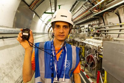 A young man wearing safety equipment shows the time in a chronometer in front of a tunnel with technological equipment at CERN