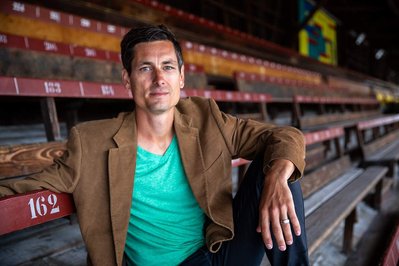 A man with a green t-shirt and brown jacket sitting in an old stadium 