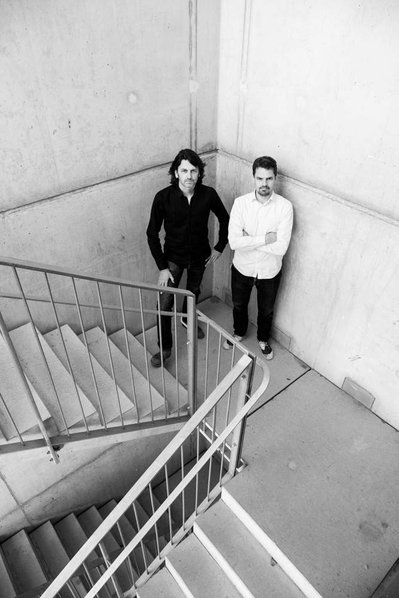 Two men, one in black outfit and the other with a white shirt look up from a staircase 