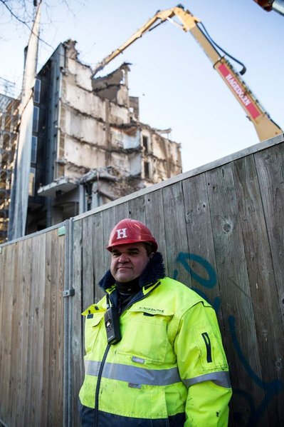 A construction worker leans on a wooden wall in front of a construction site that can be seen in the background 