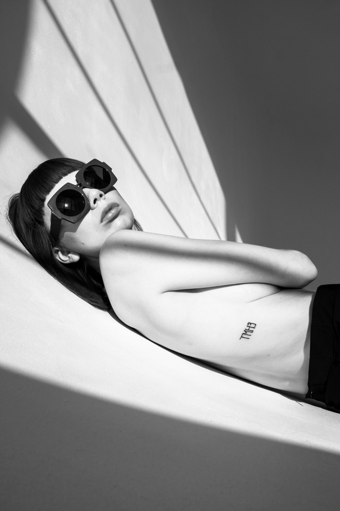 A woman with bare torso lays on a white surface wearing sunglasses.
