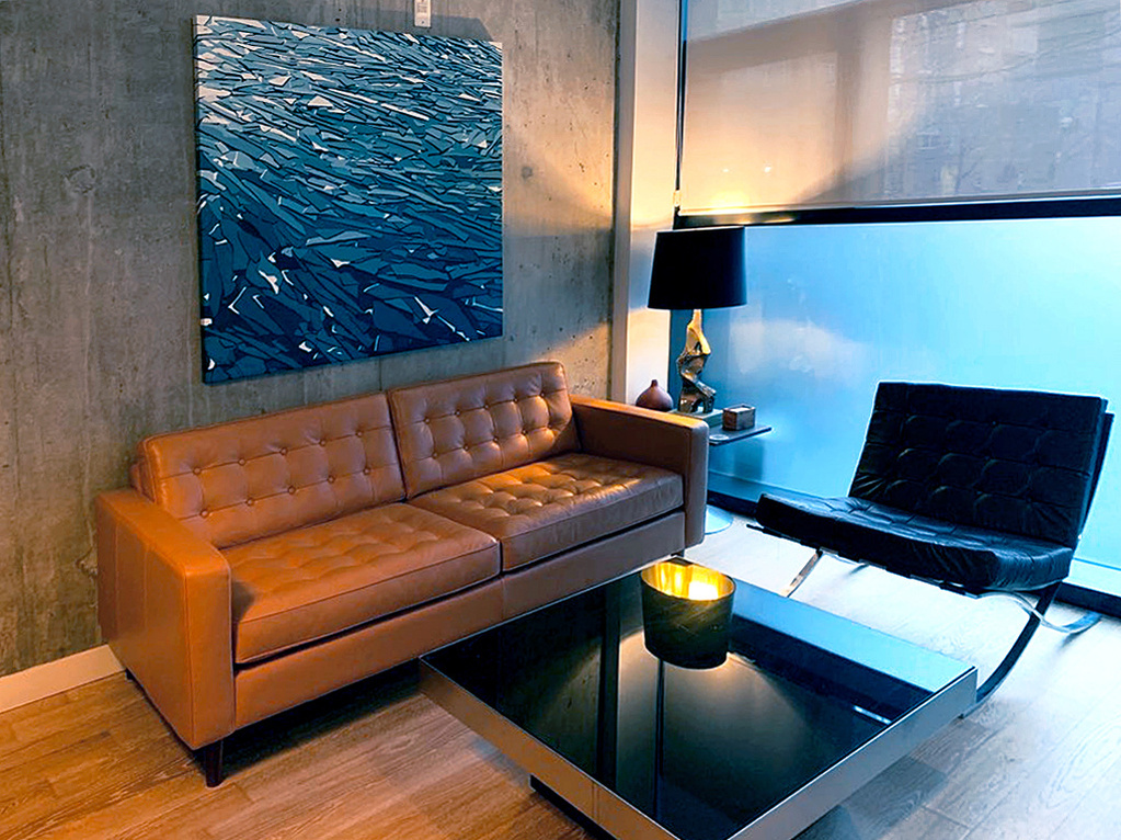 A blue painting of ice shards hangs on a concrete wall above a warm brown leather couch. 