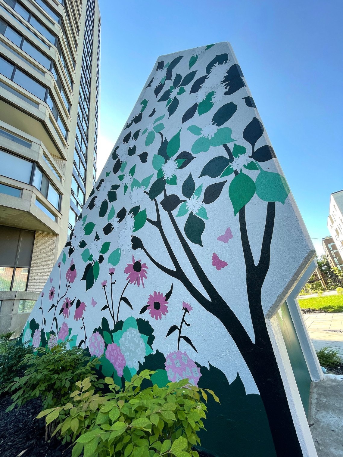 A concrete structure sits in a yard, disguised by a painted mural of plants found in the garden. A blossoming dogwood tree is the main focus with pink hydrangea and cone flowers growing around it. 