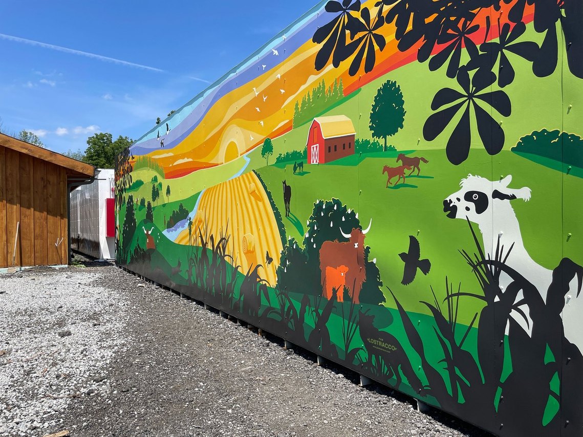 A mural shows an alpaca on the right side looking towards a farm scene of running horses, highland cows, and a corn field. 