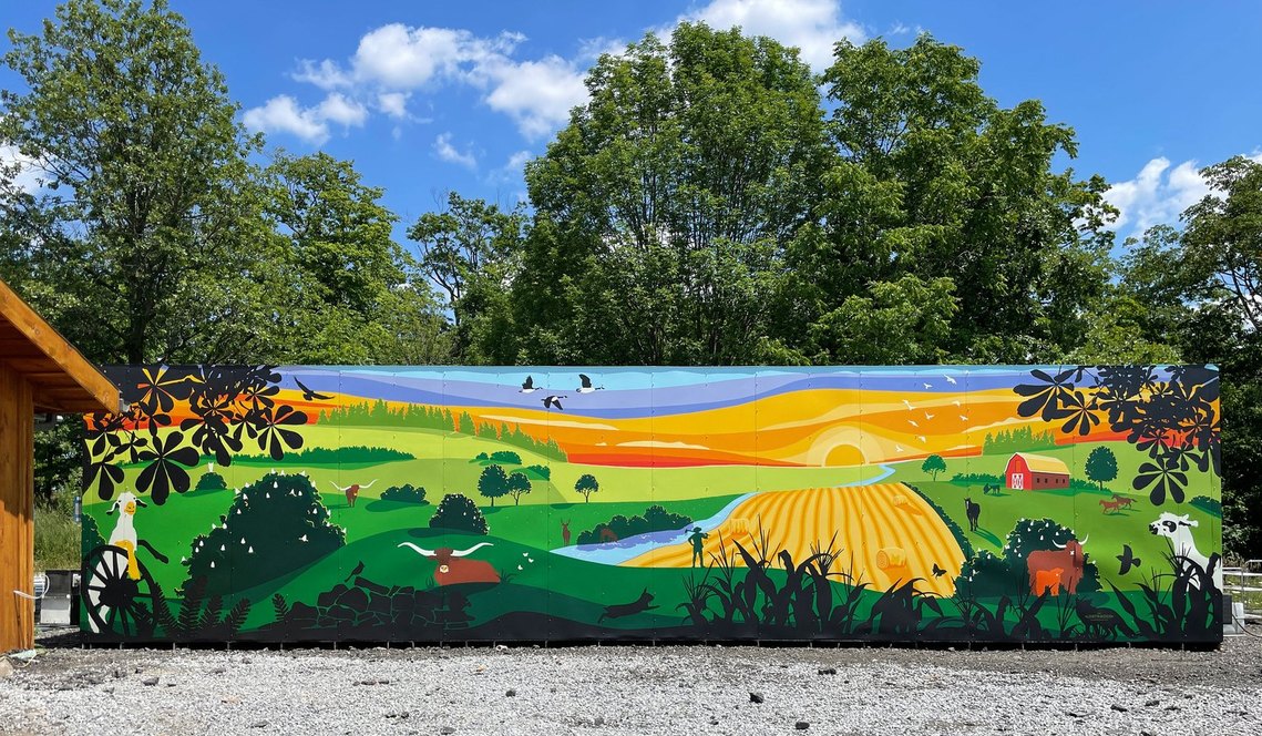 Mural on the side of a large container outside of a barn in Bradford. A countryside scene shows rolling hills and farm animals at magic hour that matches the blue sky and chestnut trees.