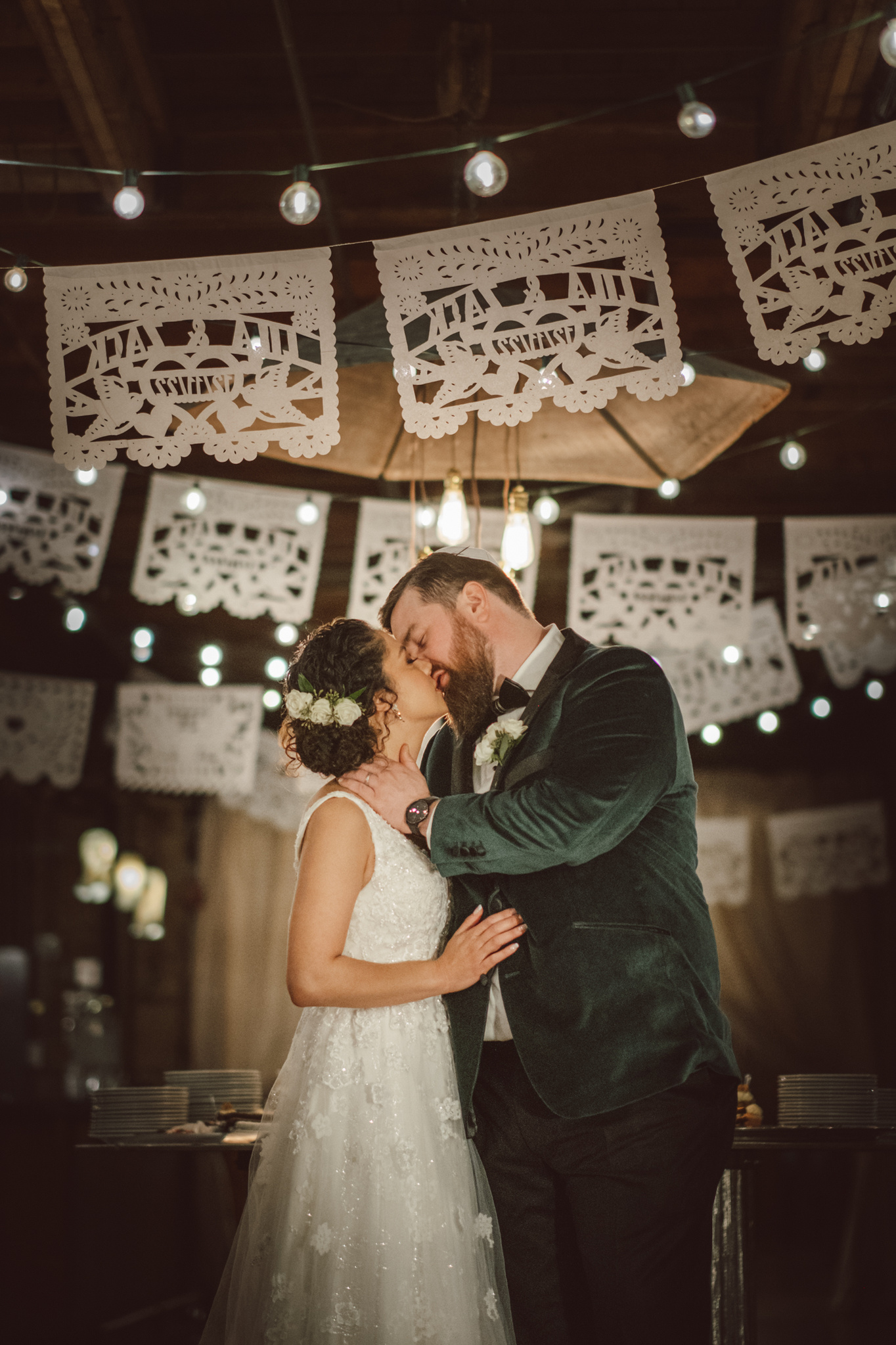 Couple kisses on their wedding day under a banner with their names and date of wedding.  Groom is wearing a green suit, Bride is wearing a white dress with flowers in her hair.  Portrait orientation. Elgin, IL wedding at The Haight.