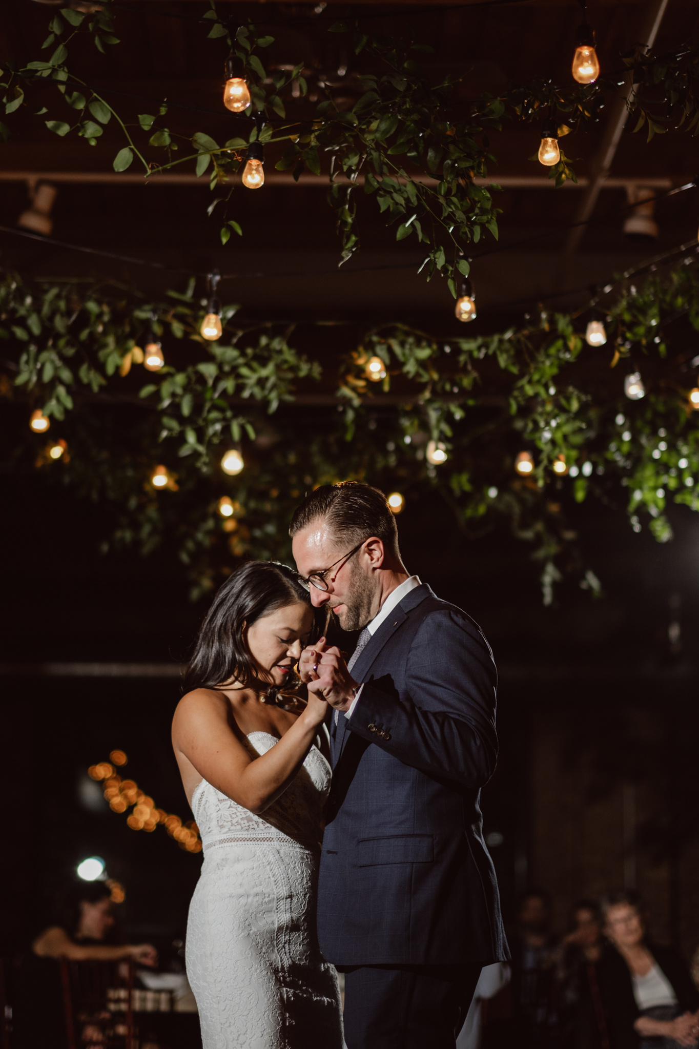 Wedding couple dances under café lights and greenery at City View Loft Chicago.  First Dance, couple holds hands as they slow dance.