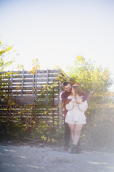 Milwaukee engagement photography session in the Riverwest neighborhood.  Couple embraces in front of a fence with the sun behind them, backlighting the scene.