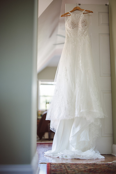 white wedding dress hanging on door during bridal prep of wedding day.  color photo, dress is on a wooden hanger hanging longways.
