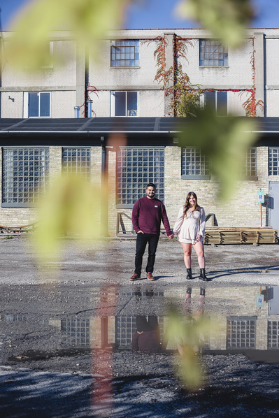 Milwaukee engagement photography session in the Riverwest neighborhood.  Couple holding hands with changing leaves in the foreground with industrial building behind them.  Amorphic Beer in background.