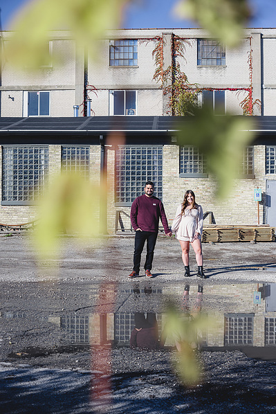 Milwaukee engagement photography session in the Riverwest neighborhood.  Couple holding hands with changing leaves in the foreground with industrial building behind them.  Amorphic Beer in background.