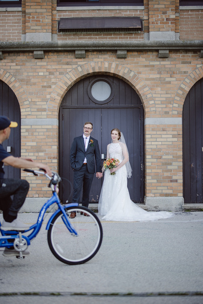 bride and groom stand in front of a doorway on their wedding day in milwaukee wisconsin at south shore terrace.  a man on a bicycle rides through the frame in the foreground, with the bride and groom looking at him.