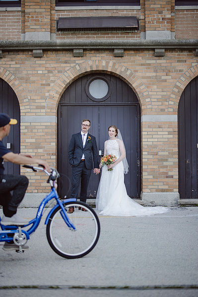 bride and groom stand in front of a doorway on their wedding day in milwaukee wisconsin at south shore terrace.  a man on a bicycle rides through the frame in the foreground, with the bride and groom looking at him.