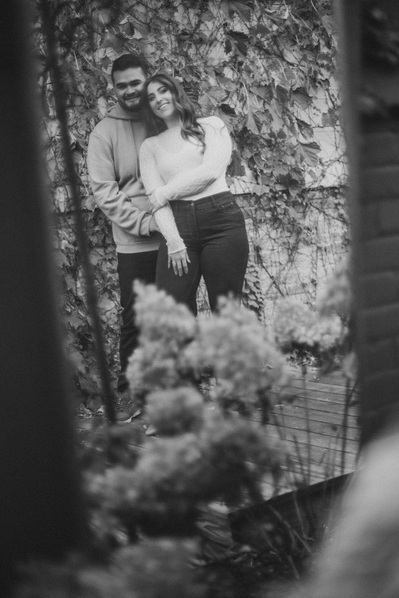 Milwaukee engagement photography session in the Riverwest neighborhood.  At Amorphic Beer in Beer Alley, the couple is embracing and reflected in a mirror with plants all around.  Photograph is in black and white.
