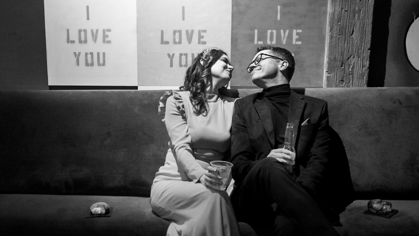 black and white photograph.  wedding couple sits on couch under sign that says 