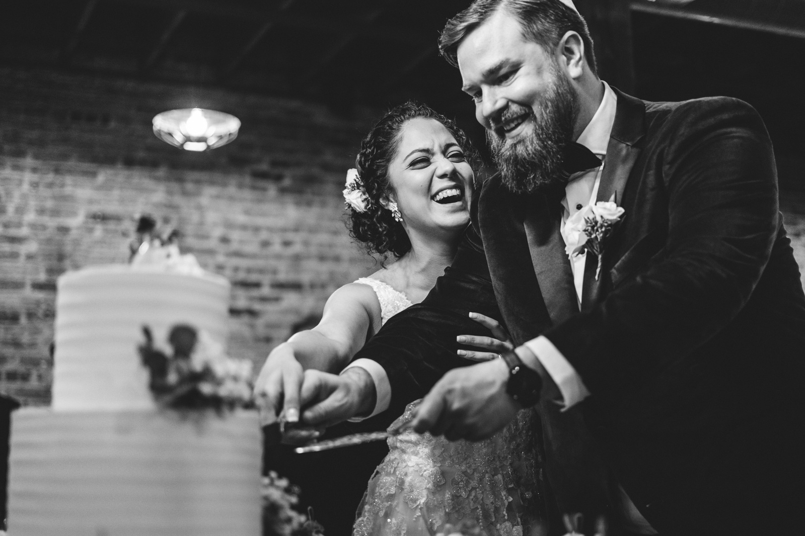 Black and white photograph of couple cutting the cake while smiling and laughing.  Groom wears a yamulke and Bride has flowers in her hair.  Cake is white with lego figurines on top.