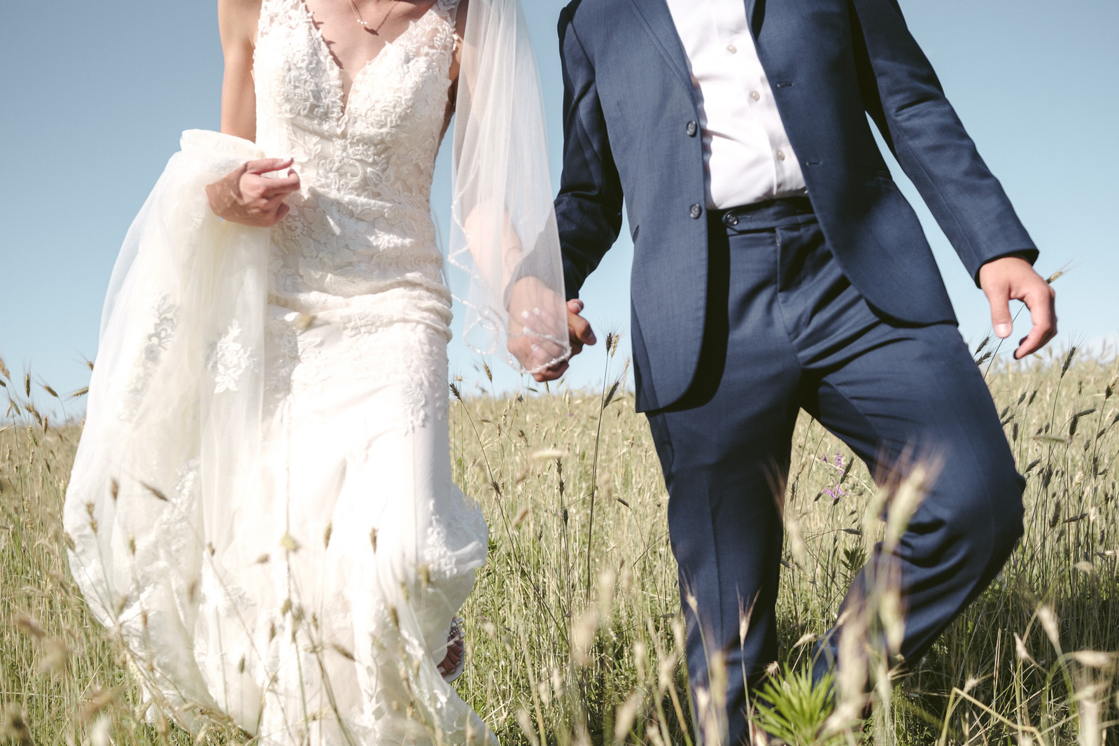 couple walks hand in hand through a field of tall grass on their wedding day.  white wedding dress, navy blue suit.  Green grass and blue sky