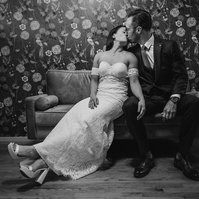 Couple sits on couch, kissing, in black and white.  Wedding day, bride is in white dress and groom is in dark suit.  flowered wallpaper is behind them.