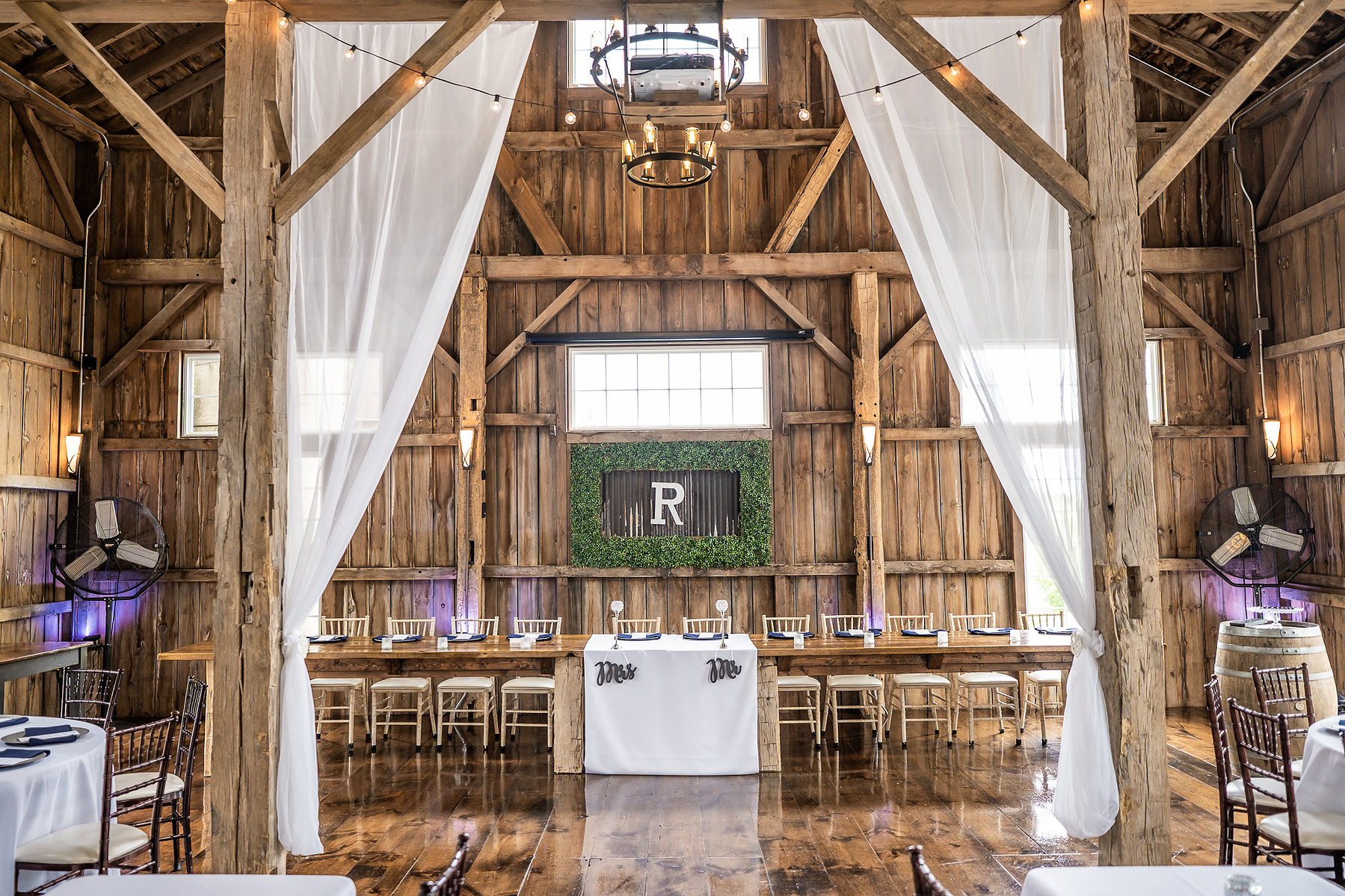 Cupola Barn in Oconomowoc, Wisconsin as one of the top barn wedding venues in Milwaukee.  Photograph is of  the inside of the barn, facing the head table with drapery, wood, and café lights in the frame along with a chandelier.