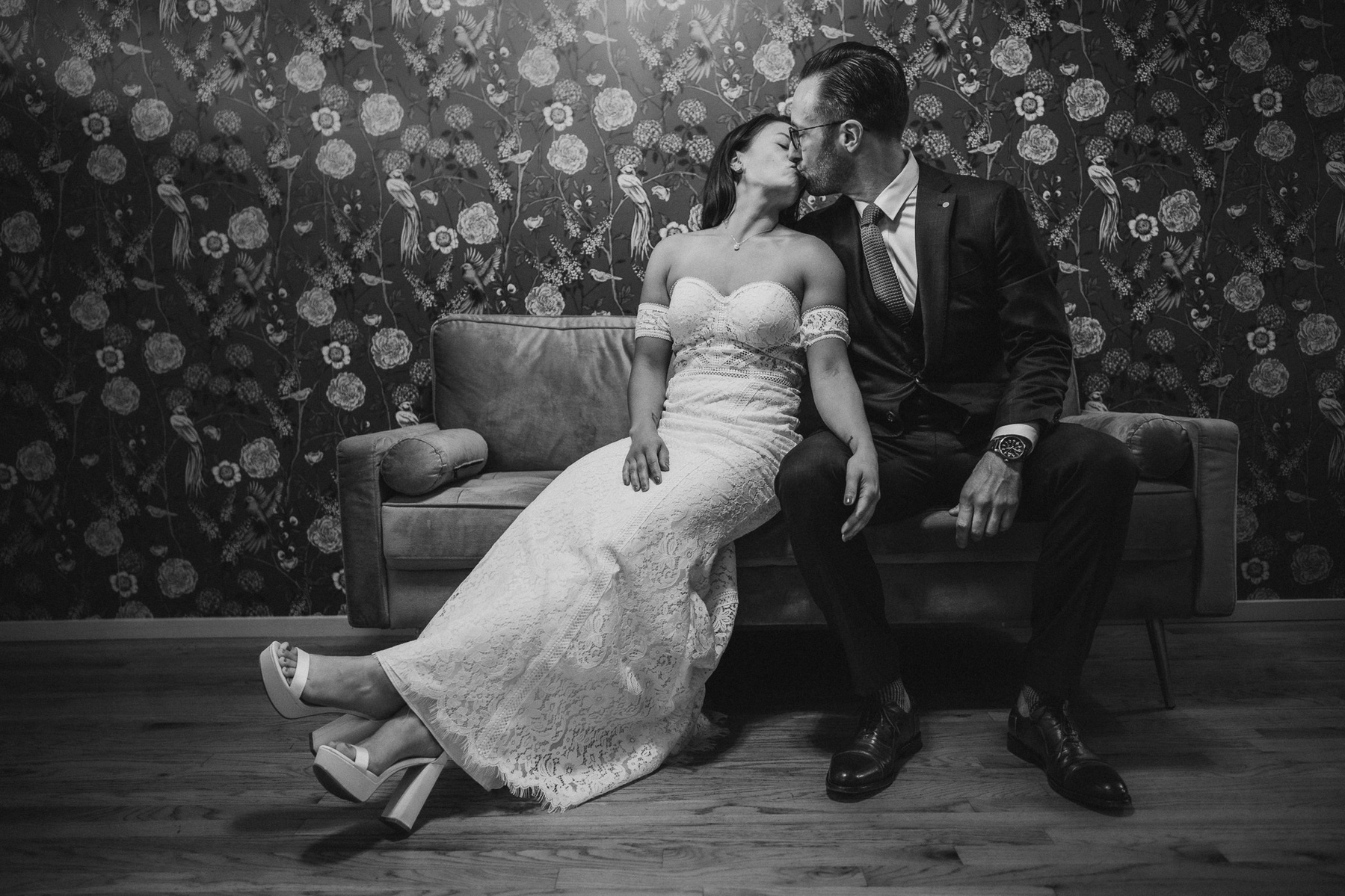Couple sits on couch, kissing, in black and white.  Wedding day, bride is in white dress and groom is in dark suit.  flowered wallpaper is behind them.