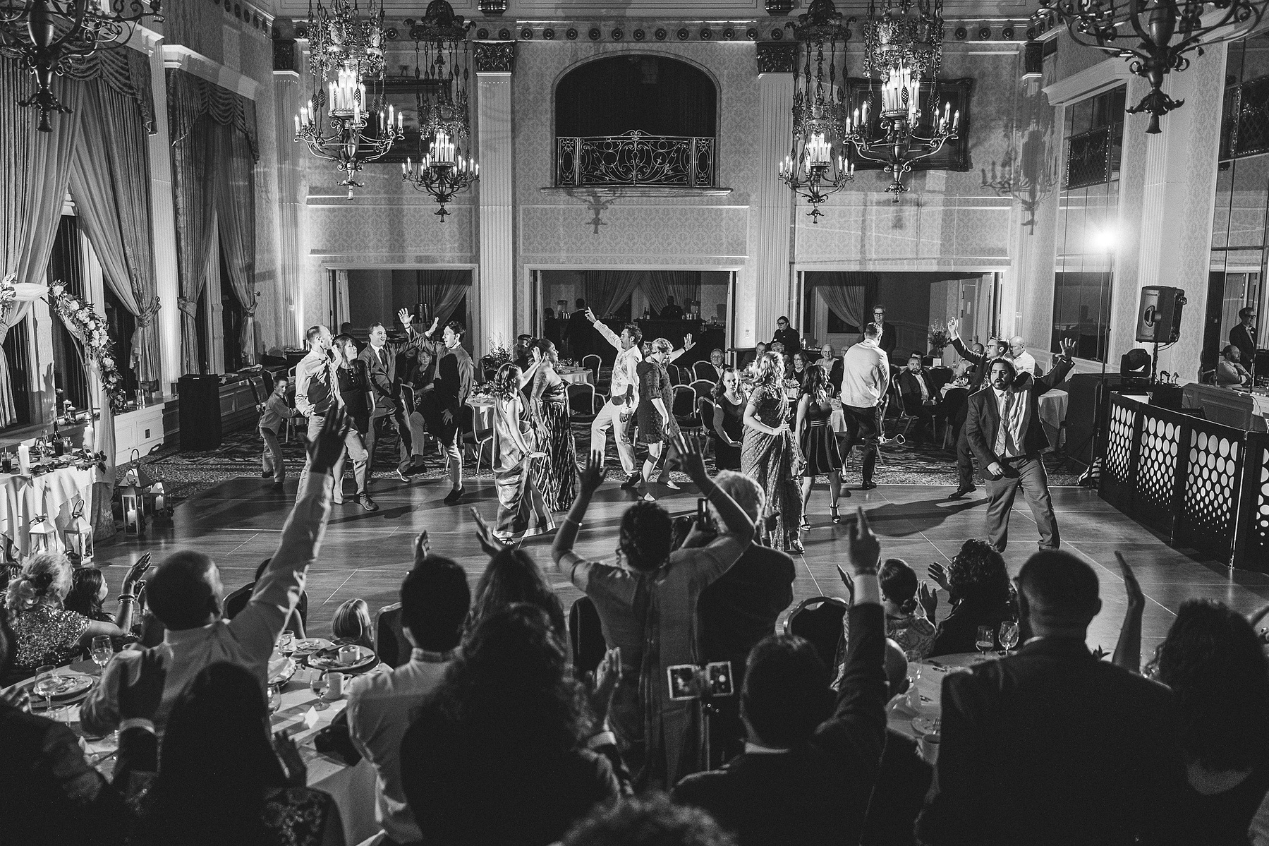 The Pfister as one of the top wedding venues in Milwaukee, Wisconsin.  Photograph is black and white dancefloor shot inside of the ballroom.  Several guests dancing to a choreographed routine during an Indian wedding while other guests look on.