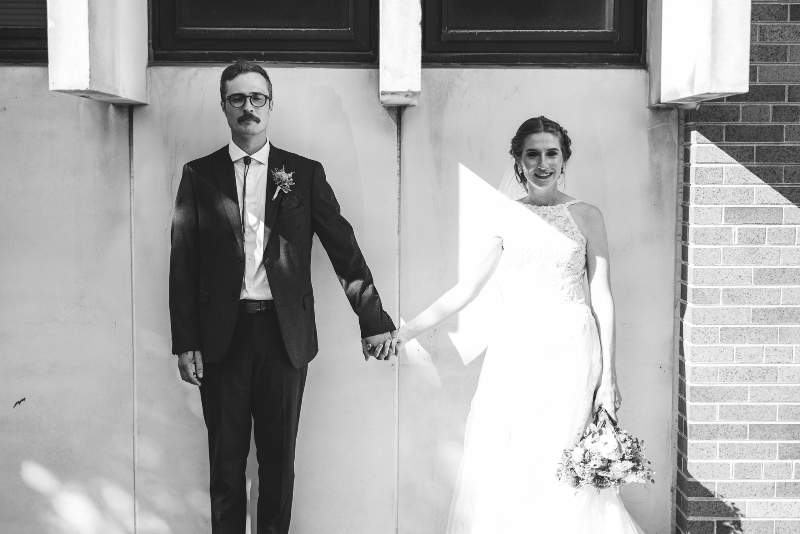 Milwaukee wedding with Bride and Groom portrait standing in front of wall with interesting architecture.  white wedding dress, dark grey suit.  Couple is holding hands and there are streaks of light shining across them.
