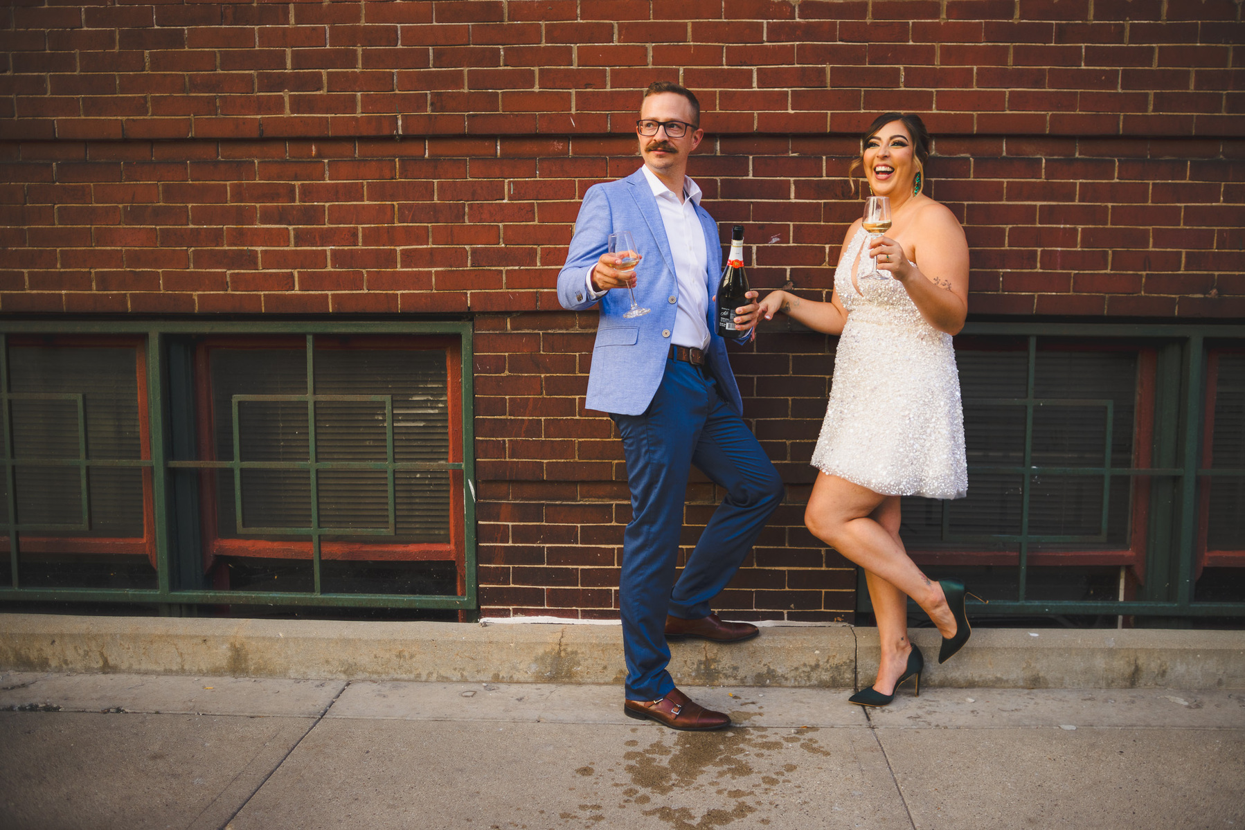 Milwaukee engagement shoot, outdoors, color, couple stands against brick wall drinking champagne