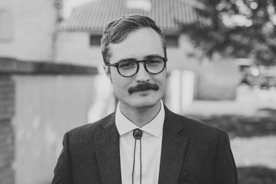 Medium shot of groom just before first look with bride in Milwaukee Wisconsin at South Shore Terrace.  Black and white photograph, groom is wearing a bolo tie and glasses and has a mustache, smiling slightly at the camera.