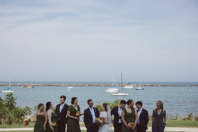 bridal party photograph in milwaukee wisconsin at south shore terrace.  Lake Michigan is in focus in the background, with sailboats on the water.  the bridal party including the bride and groom are in the foreground of the shot, out of focus, conversing.