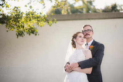 bride and groom stand in front of a white wall and under a tree on their wedding day at south shore terrace in milwaukee, wisconsin.  the bride is looking to the right of the frame while the groom is looking to the left of the frame.