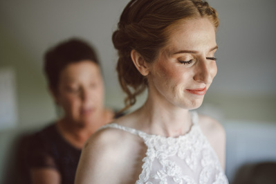 Bride looks out the window as the mother of the bride buttons the back of her dress in Milwaukee, Wisconsin on her wedding day.