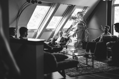 Groom sits on a chair in his AirBnB with his groomsmen around him.  Wide shot of the room in Milwaukee, Wisconsin.  Black and white photograph with long windows letting in lots of natural light with houseplants all around.