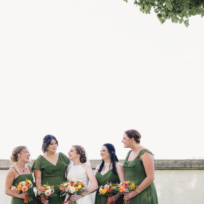 bride and bridesmaids during the wedding day in milwaukee wisconsin at south shore park.  bridesmaids are wearing green dresses which match the green leaves of a tree poking out near the top right corner of the frame.  Bride is smiling at her bridesmaids.