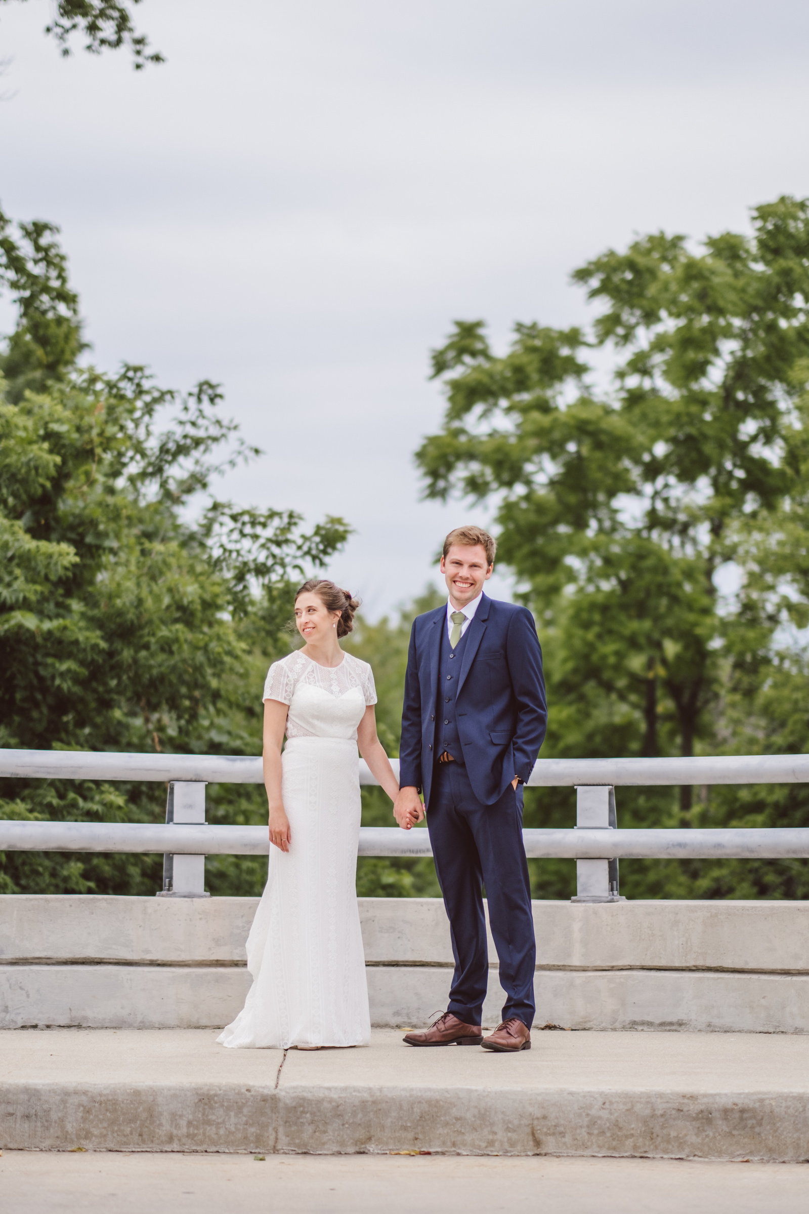 Couple hold hands on their wedding day in Milwaukee, WI at The Urban Ecology Center.  Portrait orientation, groom in blue suit, bride in white dress stand on a bridge.