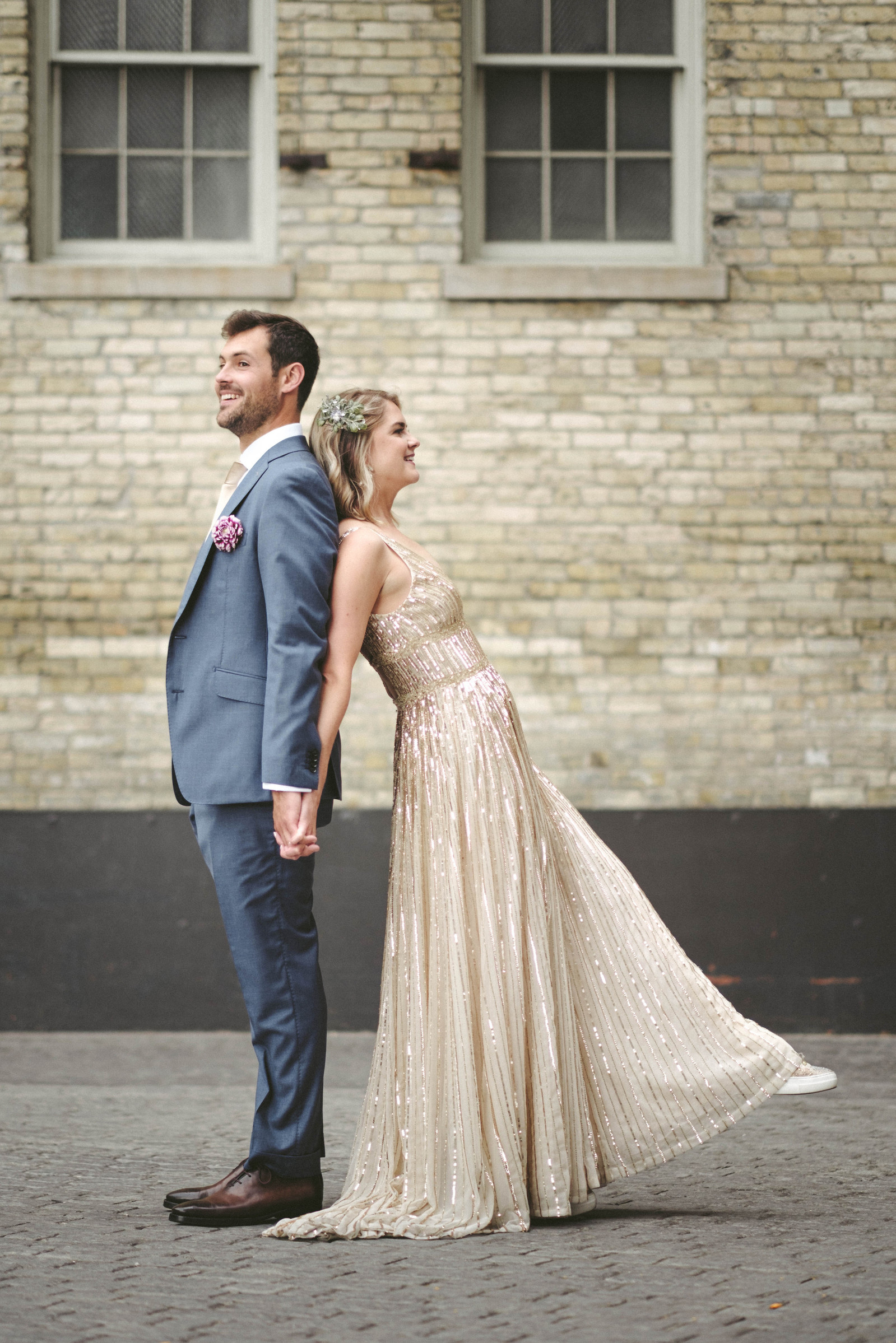 Couple stand back to back on their wedding day in Milwaukee, Groom in blue suit, smiling and looking off into the distance.  Bride is leaning her back against the groom and has her leg extended out in front.  Standing in front of cream colored brick wall.